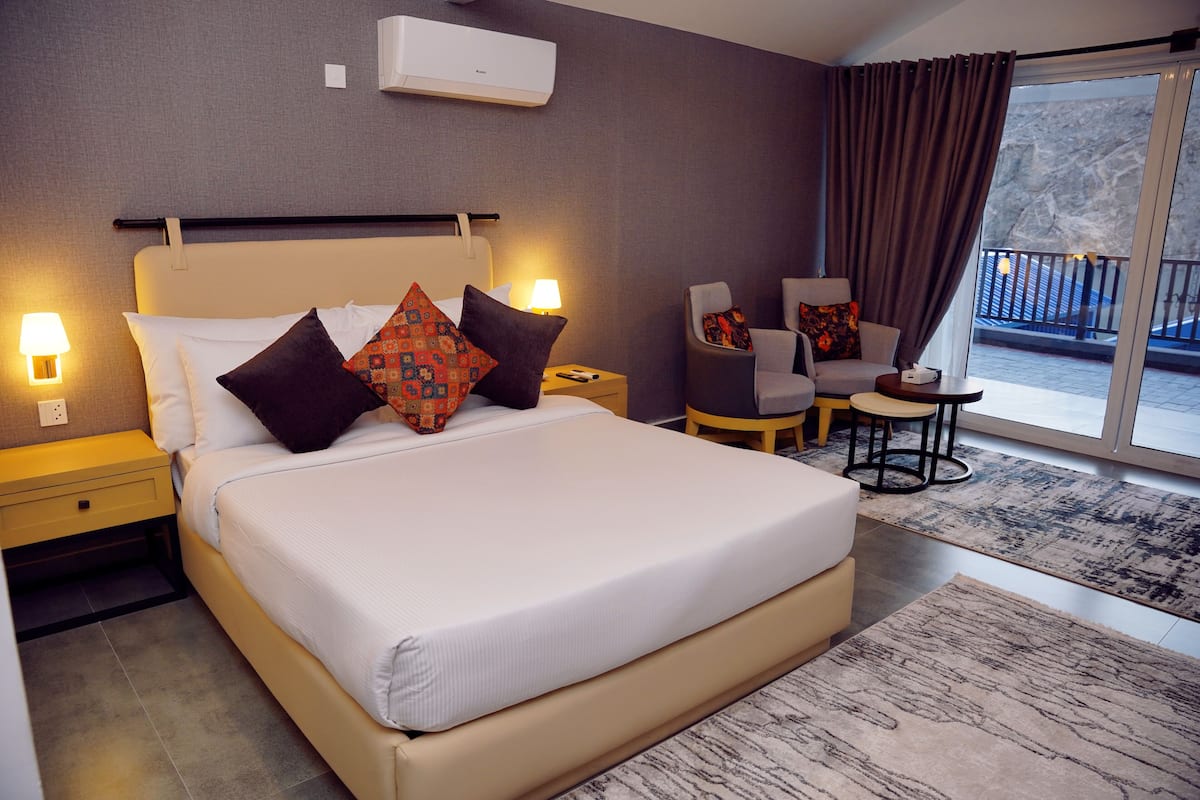 A modern hotel room with a large bed, two bedside tables, a chair, a small table, and a balcony with a view.