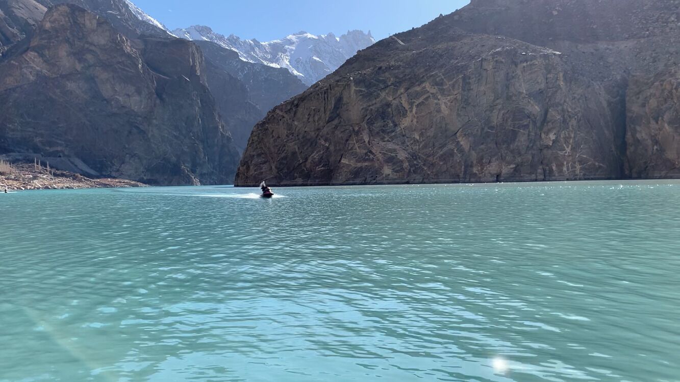 A calm lake with a person on a jet ski, surrounded by rocky mountains and snow-capped peaks.