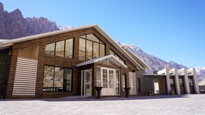 A modern building with large windows and a sloped roof, set against a mountainous backdrop. - Lodges By Baron Hunza Attabad Lake (Aina Abad)