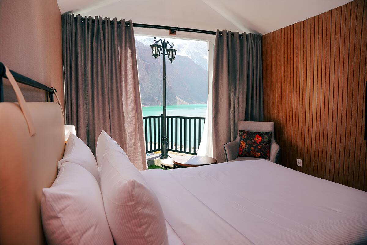 A hotel room with a large bed, a chair, a table, and a view of a mountain and lake through the window.
