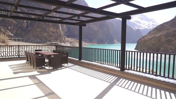 A covered outdoor seating area with a table and chairs, overlooking a turquoise body of water and mountainous terrain. - Lodges By Baron Hunza Attabad Lake (Aina Abad)