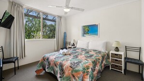 A hotel room with a bed, a ceiling fan, a TV mounted on the wall, a window with curtains, and a painting of a beach scene. - Miami Beachside Holiday Retreat (Miami)