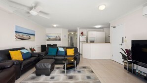 A modern living room with a black leather sofa, a glass coffee table, and a grey patterned rug. There is a flat-screen TV on the right, a ceiling fan, and a kitchen area in the background. - Miami Beachside Holiday Retreat (Miami)