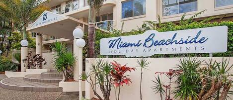 A multi-story building with a sign for Miami Beachside Holiday Apartments. There are steps leading to the entrance, surrounded by various plants and a palm tree.