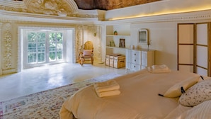 Ruime slaapkamer met een groot bed, een houten schommelstoel, een dressoir en een groot raam.
