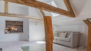 Een ruime loft met zichtbare houten balken, een glazen dak en een grijze bank met een gele voetenbank.
