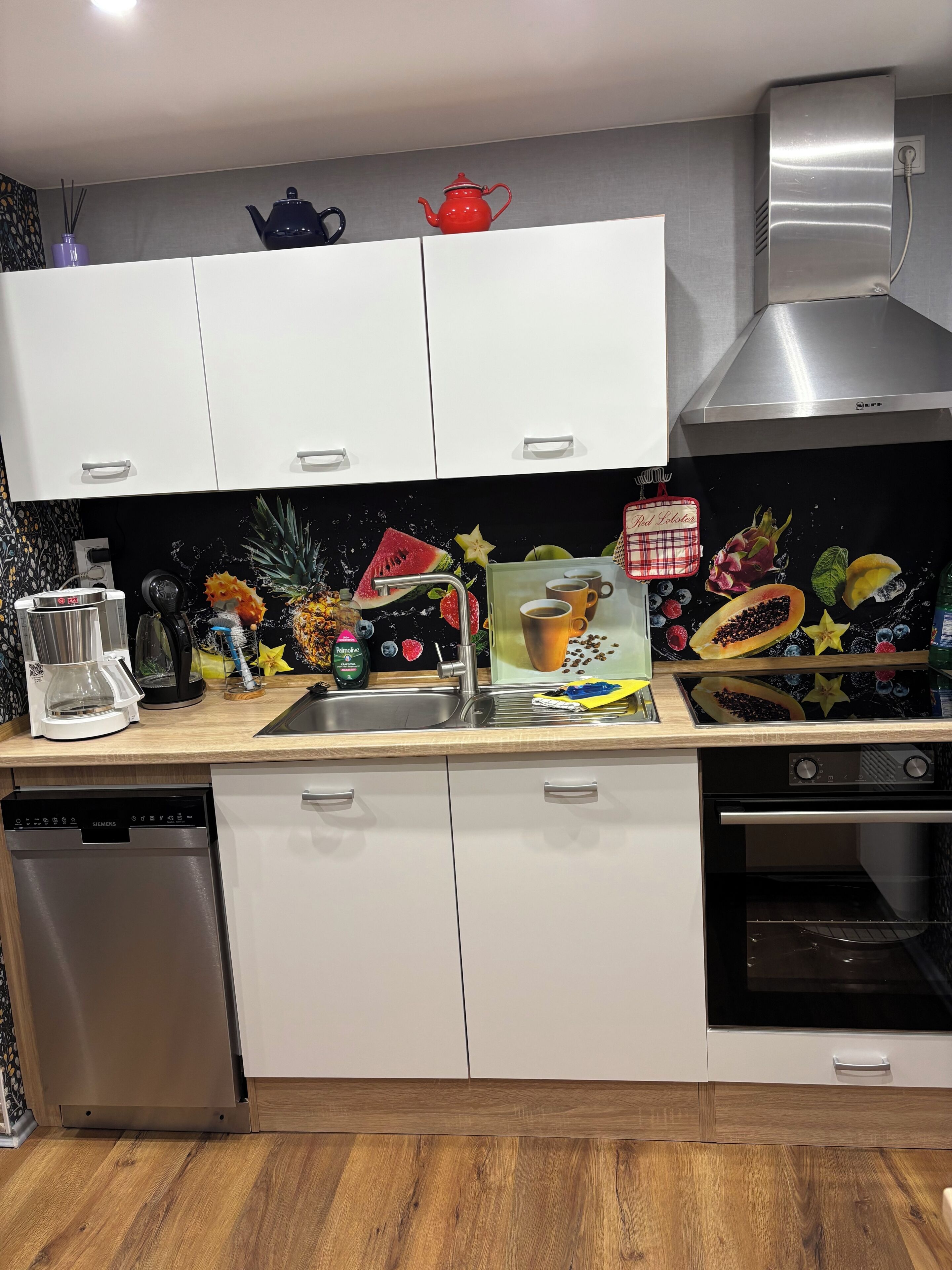 Een moderne keuken met witte kastjes, een roestvrijstalen vaatwasser, een koffiezetapparaat en een rode theepot op het aanrecht.