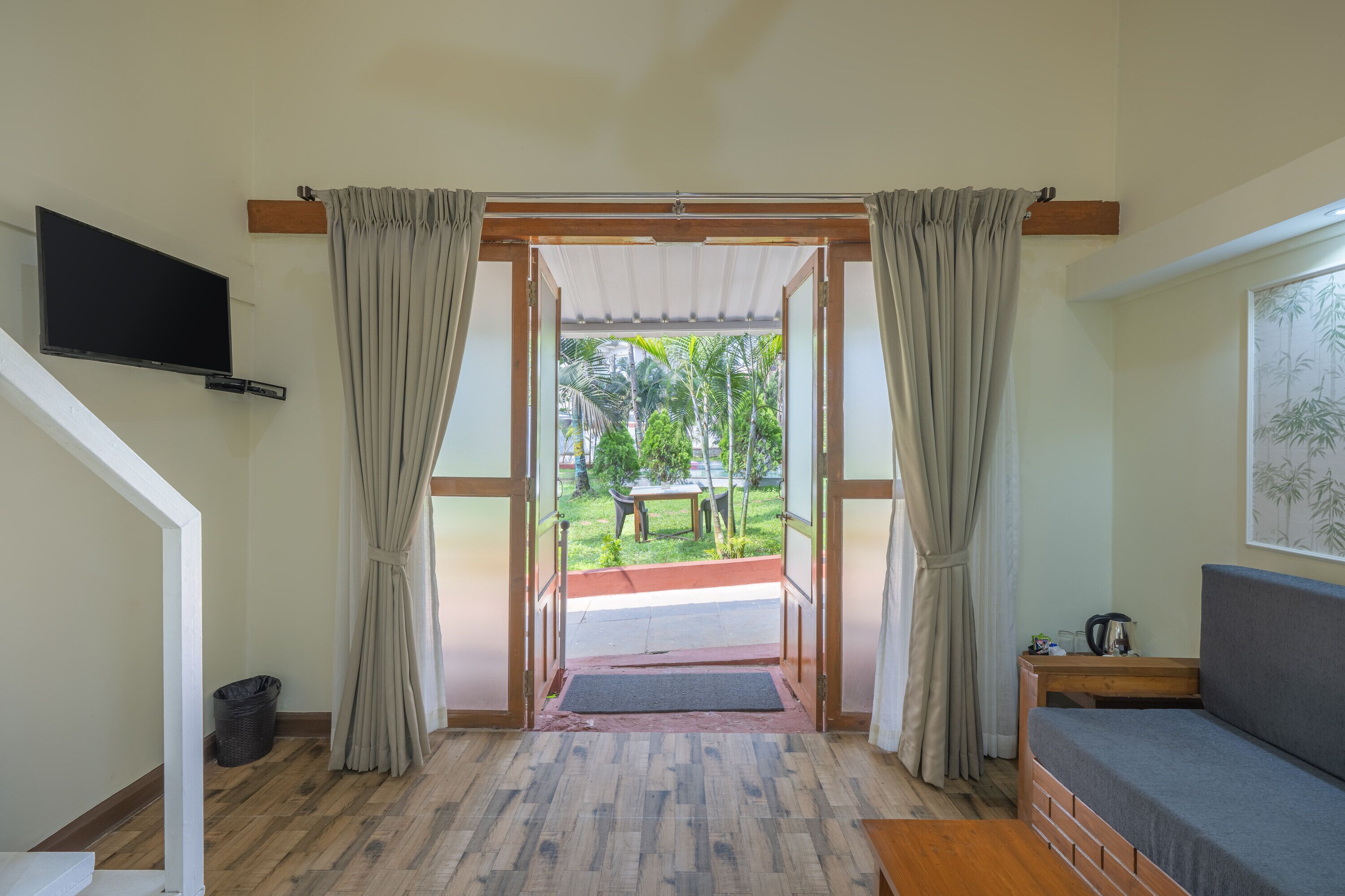 A room with a wooden floor, a sofa, a television mounted on the wall, and a view of a garden through an open door.