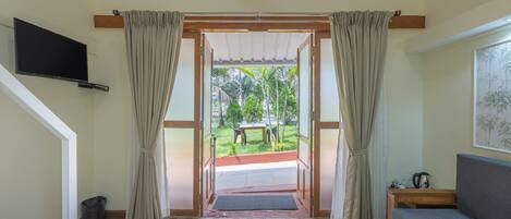 A room with a wooden floor, a sofa, a television mounted on the wall, and a view of a garden through an open door.