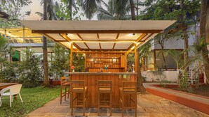 Zona de bar al aire libre con taburetes de madera y una barra, rodeada de vegetación y una zona de estar cubierta.