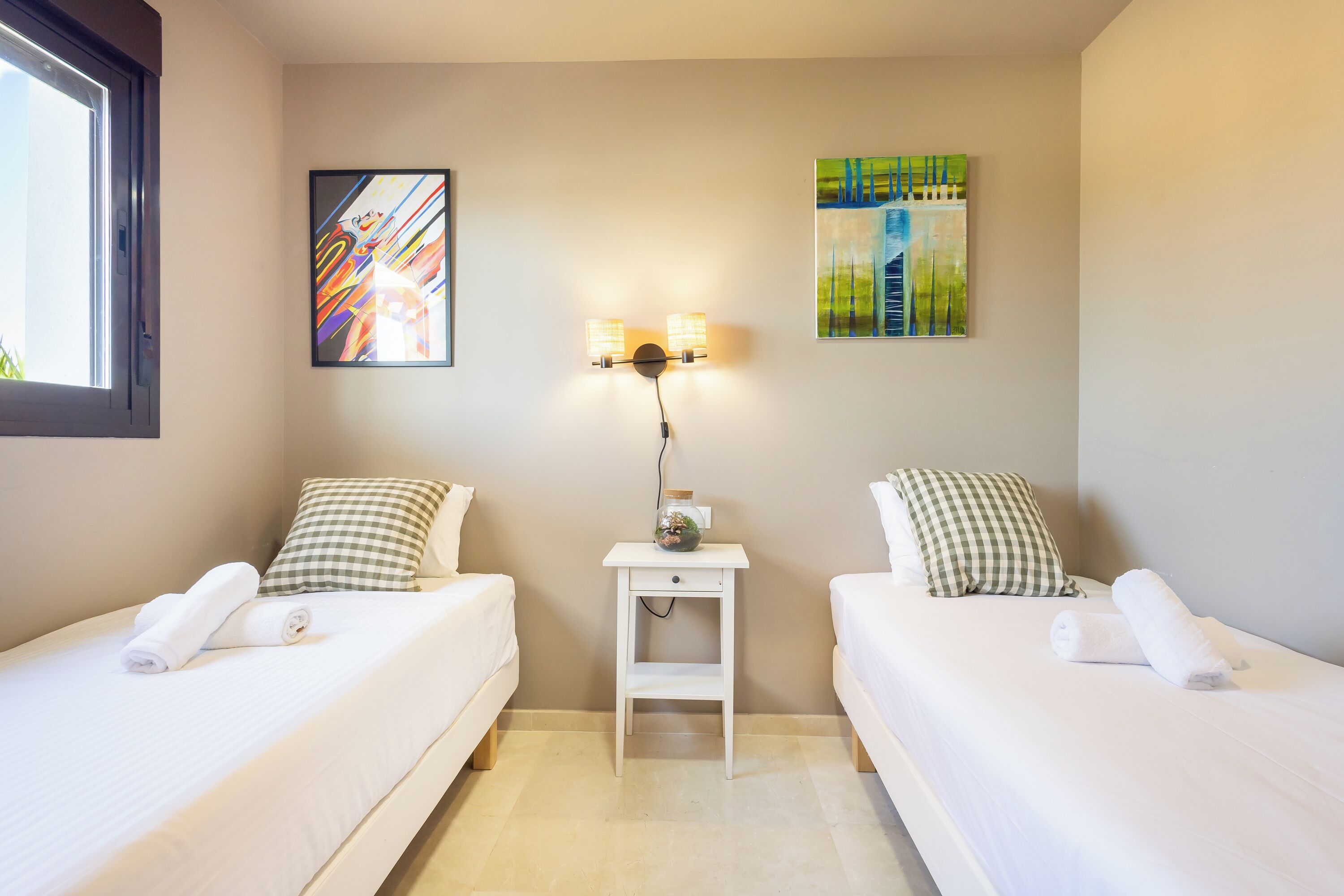 A hotel room with two beds, a nightstand with a lamp, and artwork on the walls.