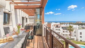Een balkon met een eettafel, een glazen afscheiding en uitzicht op zee en gebouwen.