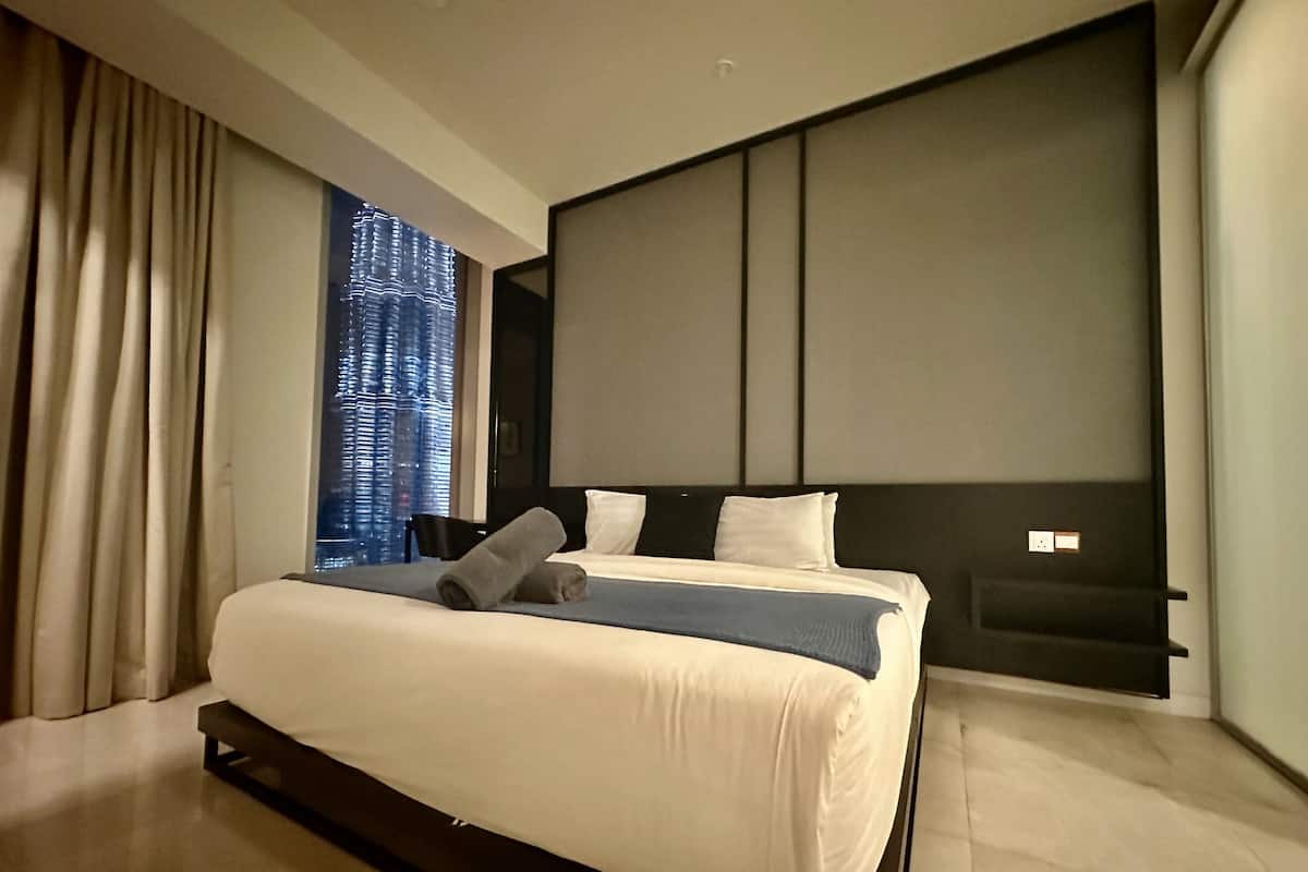 Habitación de hotel moderna con una cama grande, cabecera con iluminación integrada, vista del paisaje urbano a través de la ventana y una pared de color oscuro.