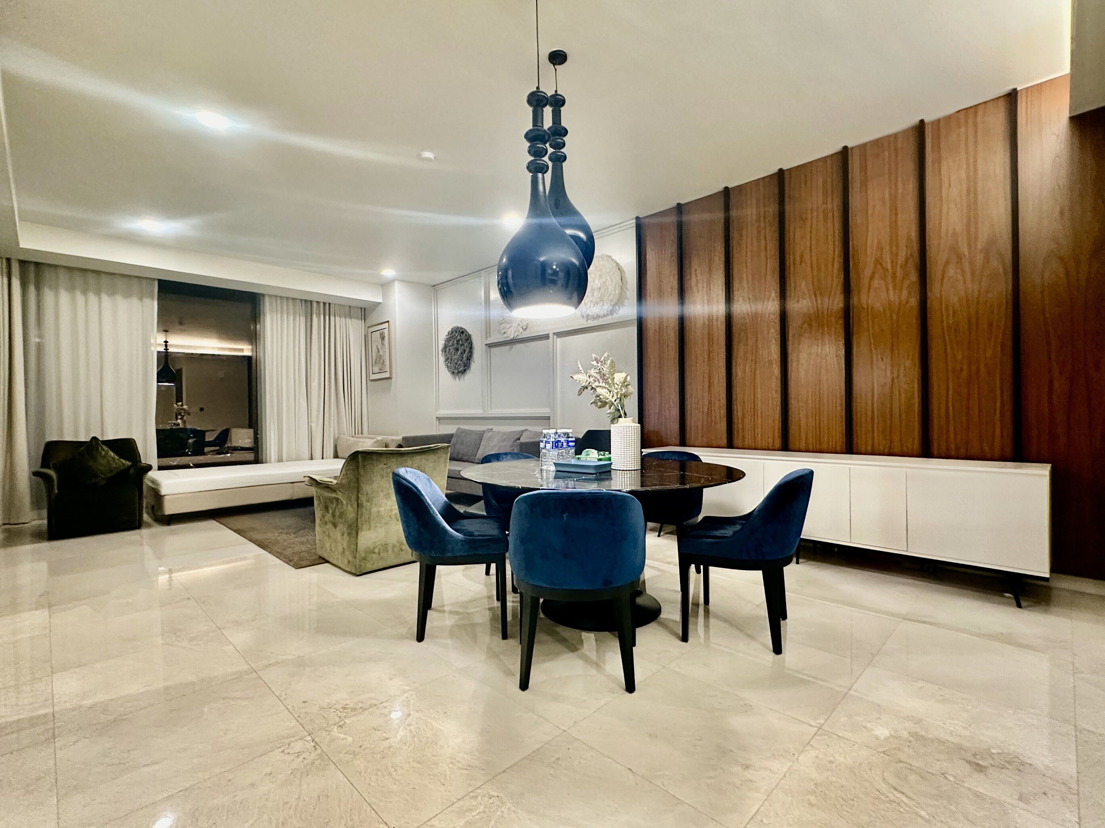 A modern living room with a round dining table, blue chairs, a large blue pendant light, and a wooden panel wall.