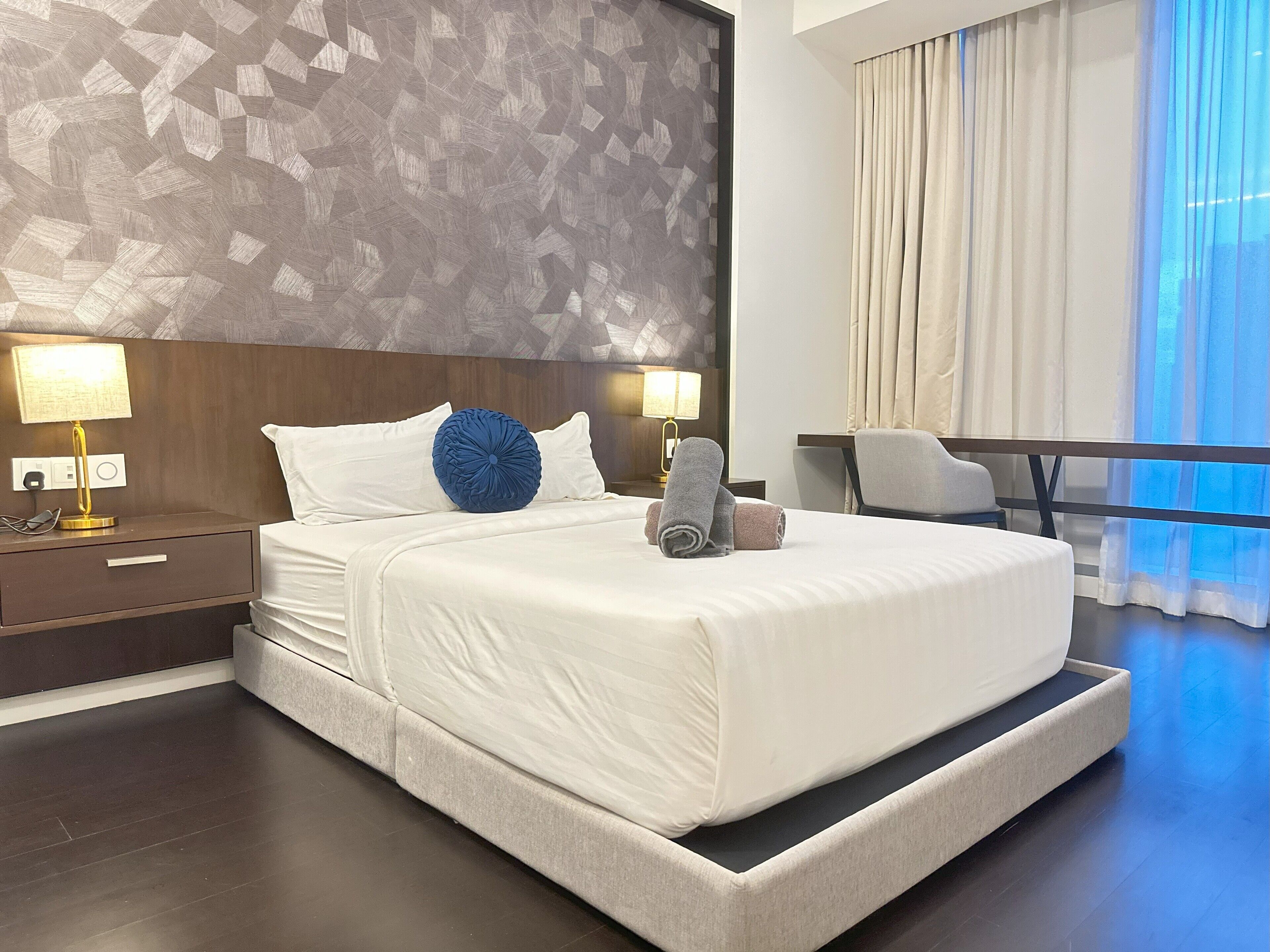 A modern hotel room with a large bed, two bedside lamps, a desk, and a view of the city through sheer curtains.