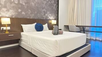 A modern hotel room with a large bed, two bedside lamps, a desk, and a view of the city through sheer curtains.