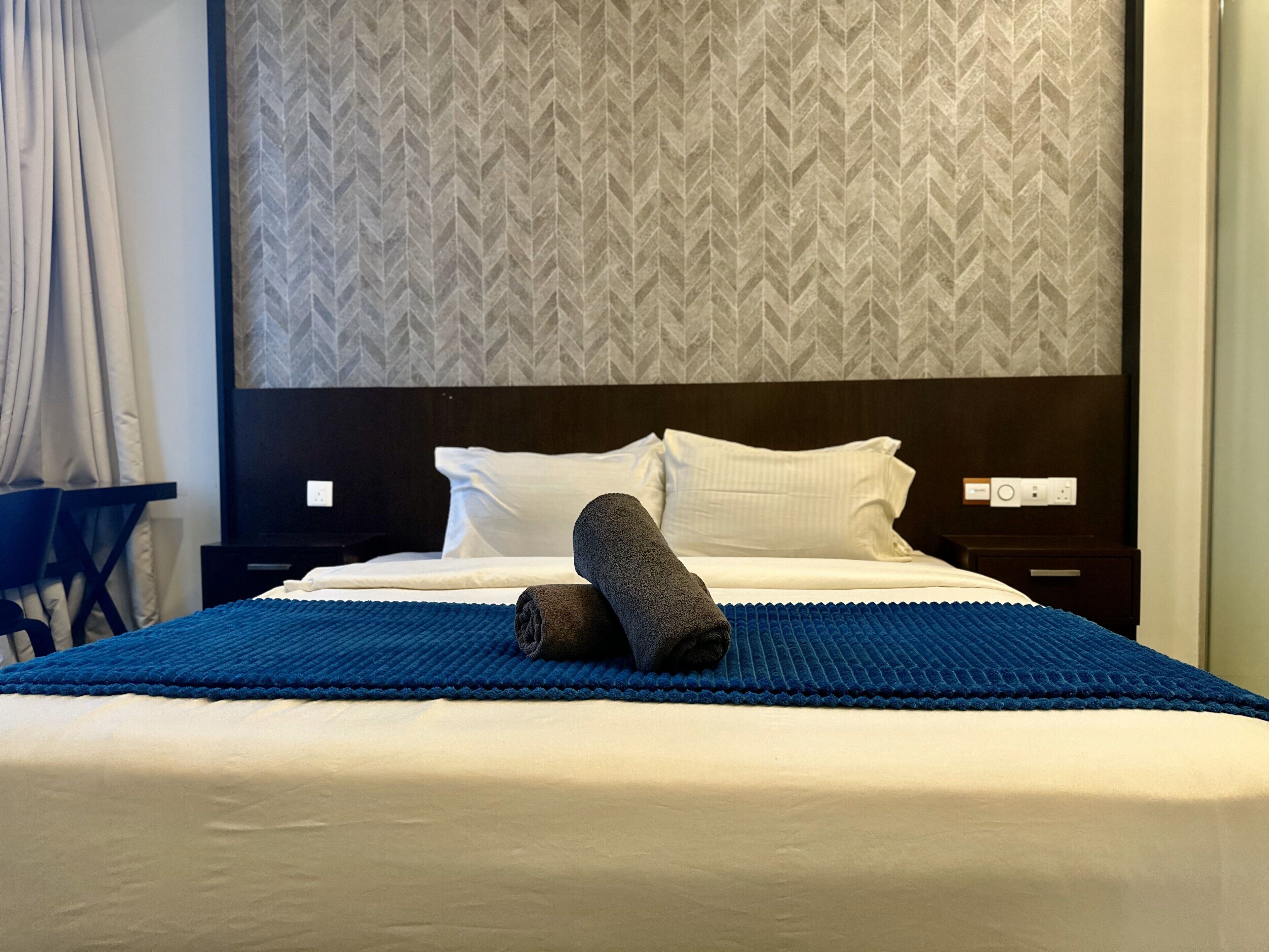 A hotel room with a bed, a blue bedspread, white pillows, and a rolled-up towel.