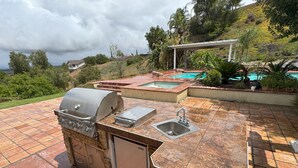 Una cocina exterior con parrilla, pileta y vistas a una piscina y colinas.
