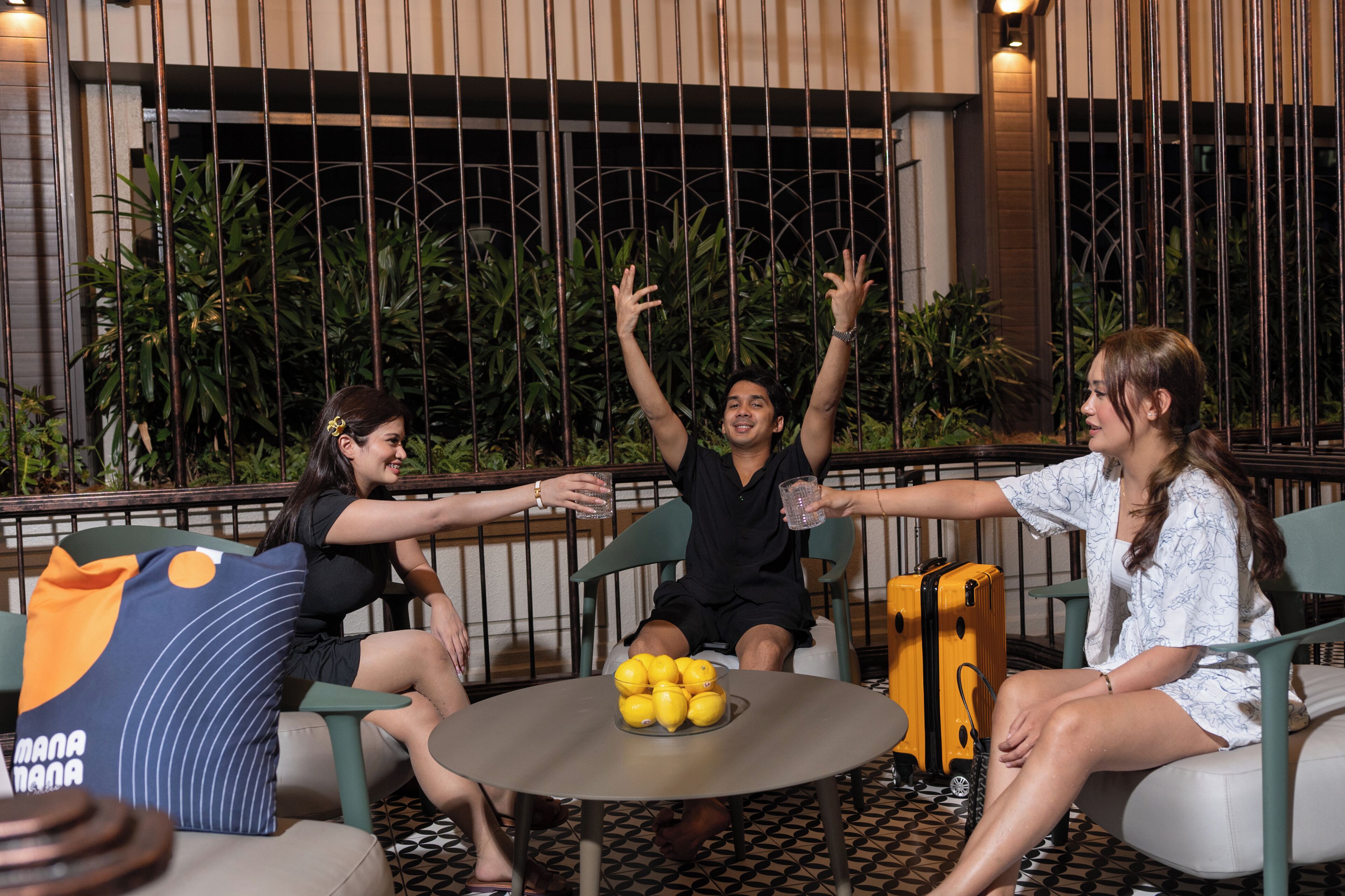 A group of people seated around a table with a bowl of lemons, a yellow suitcase, and a blue pillow with text.