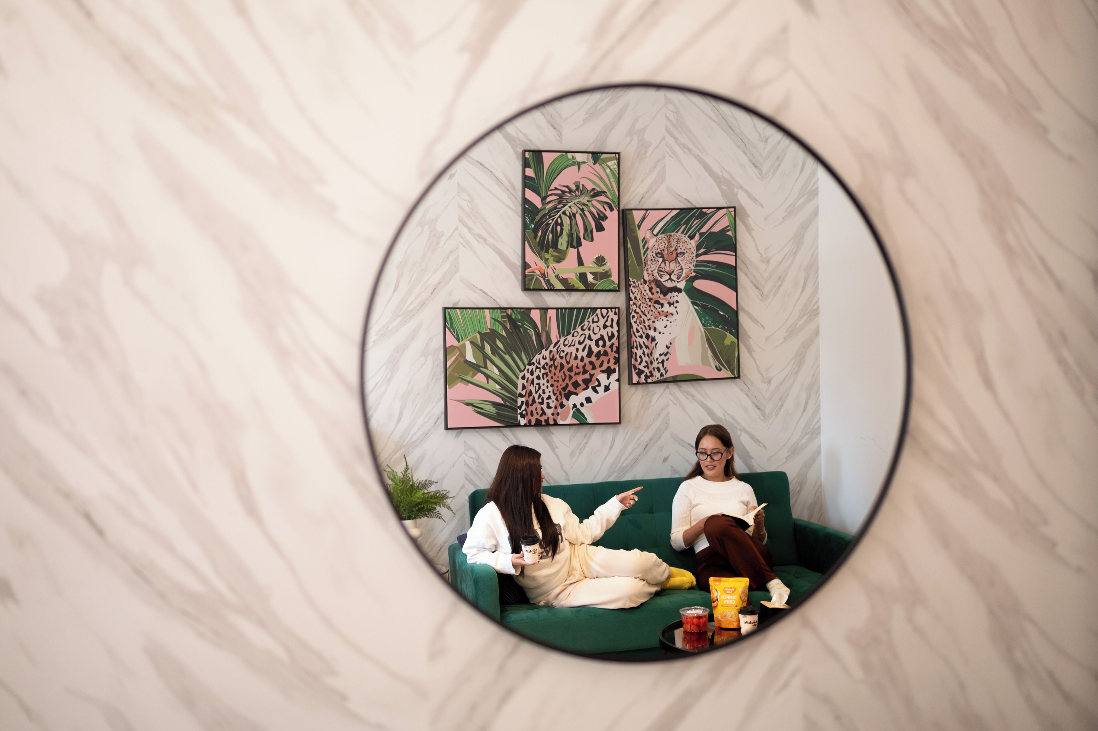 A circular mirror reflecting a room with a green sofa, two people sitting, and three leopard-themed paintings on the wall.