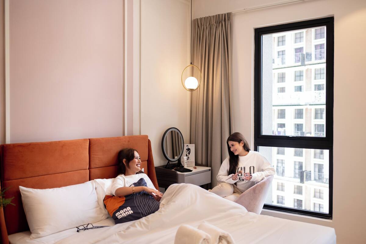 A hotel room with a large window, a bed with white bedding, a nightstand with a mirror, and a wall-mounted light fixture.