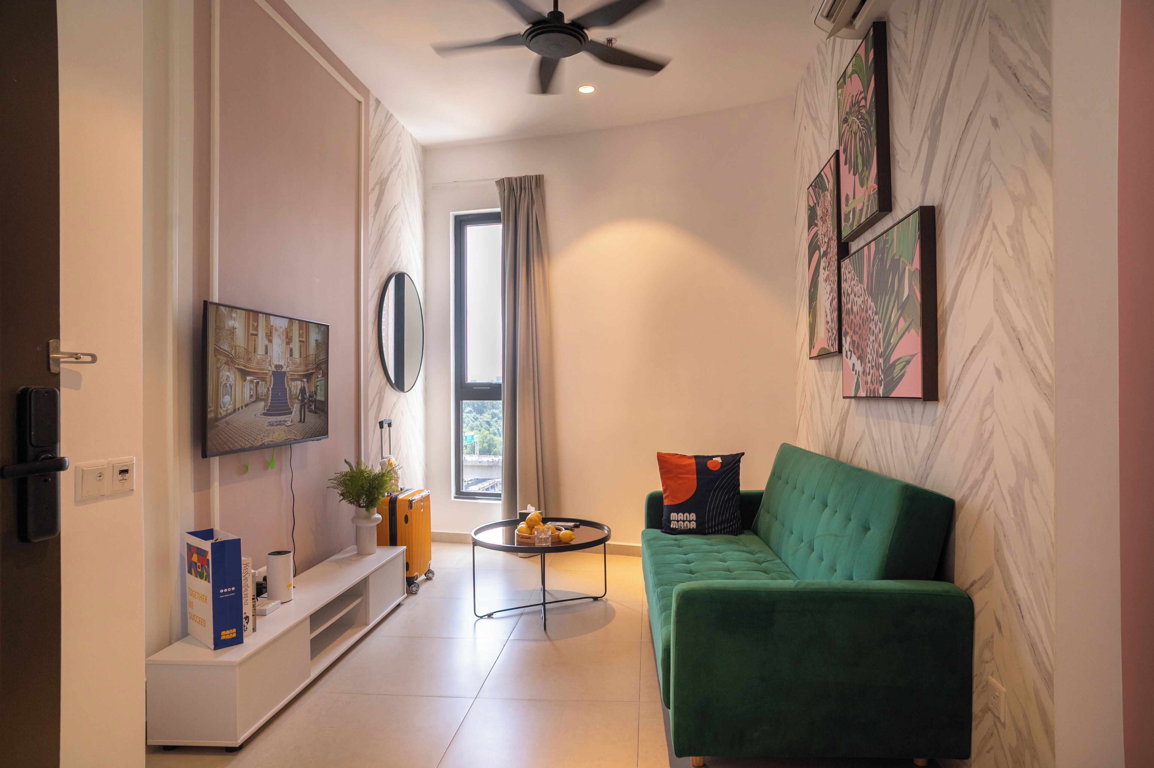 A modern living room with a green sofa, a flat-screen TV mounted on the wall, a ceiling fan, and a window with curtains.