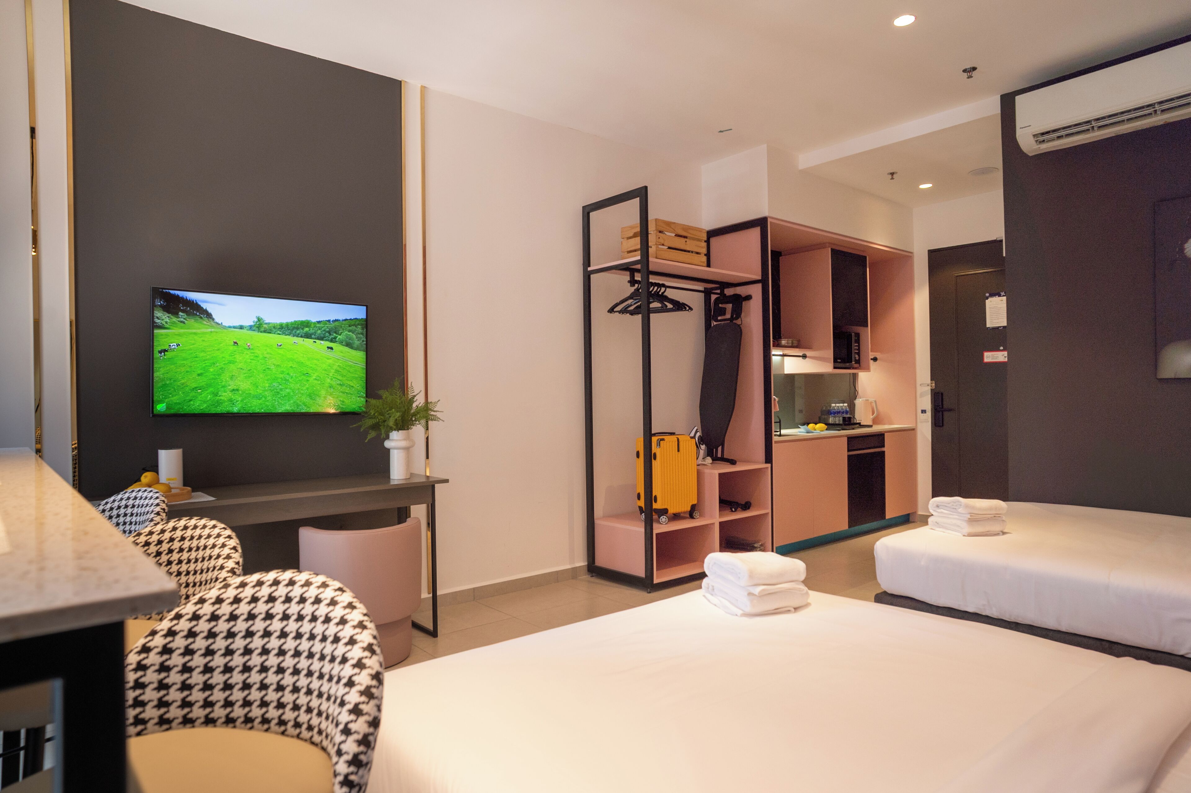 A hotel room with two beds, a TV displaying a green landscape, a desk with a plant, and a kitchenette.