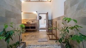 Un baño moderno con un espejo grande, un lavamanos y una zona de ducha. Hay plantas y un banco de madera en el espacio.