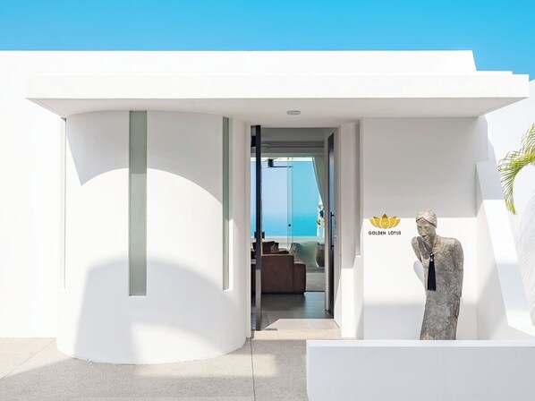 A modern building with a white exterior and a glass door leading to a room with a view of the sea. A golden lotus statue is visible on the right side.