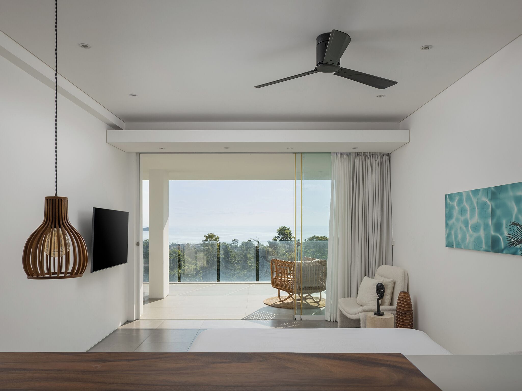 A modern living room with a large sliding glass door leading to a balcony, a ceiling fan, a mounted TV, and a piece of abstract art.