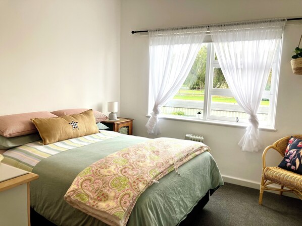 A bedroom with a bed, pillows, a bedside table with a lamp, a chair, and a window with curtains.