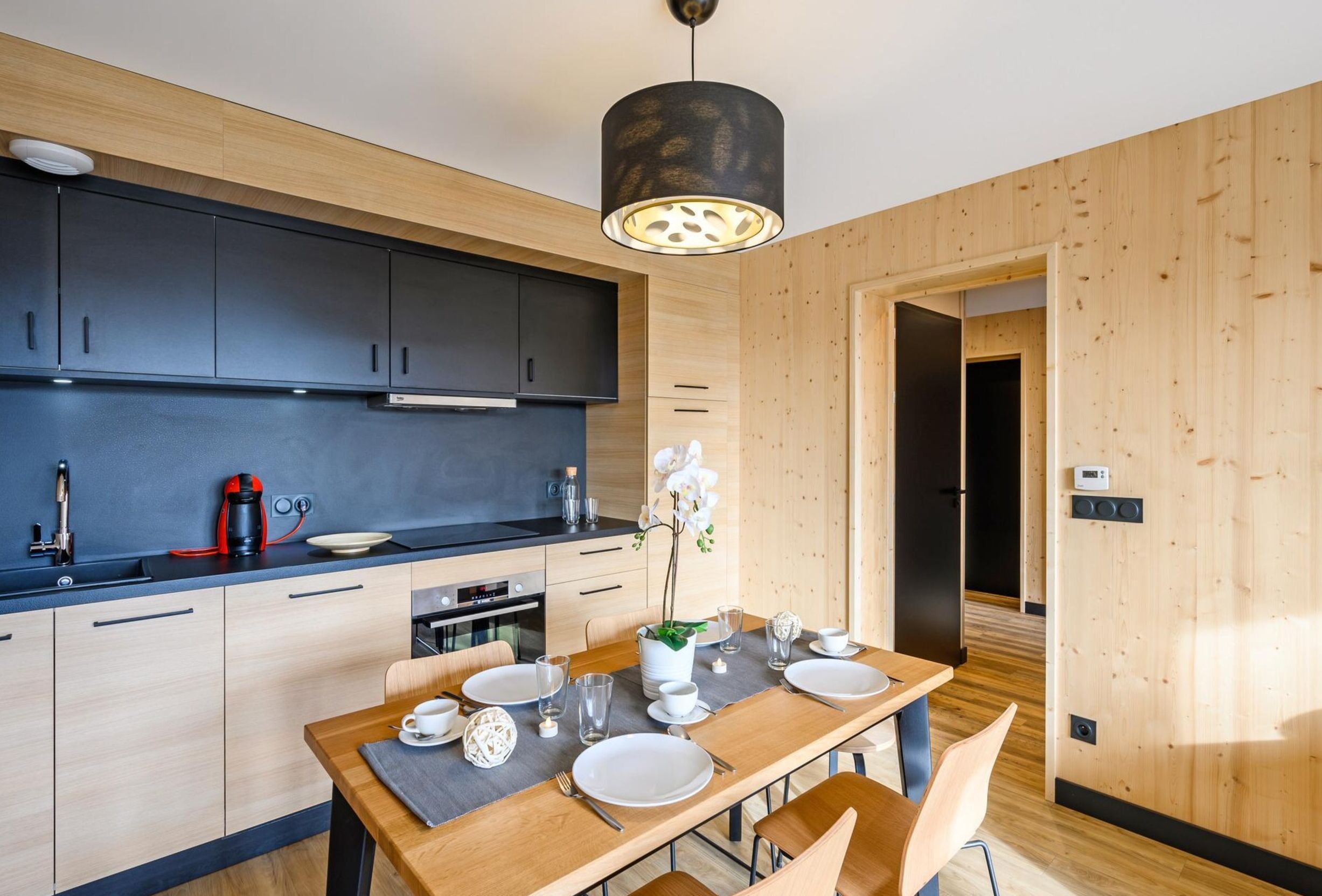 Une cuisine moderne avec des meubles de couleur sombre, une table à manger en bois pour quatre personnes et un luminaire suspendu au-dessus.