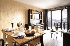 A modern dining area with a wooden table set for breakfast, a television in the background, and a balcony with a mountain view. - Apartment in Le Corbier Near Ski Slopes (Villarembert)