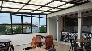 Un'ampia zona pranzo all'aperto con tavoli e sedie, ampie finestre che offrono vista su edifici e alberi, e un angolo cottura dotato di elettrodomestici moderni.