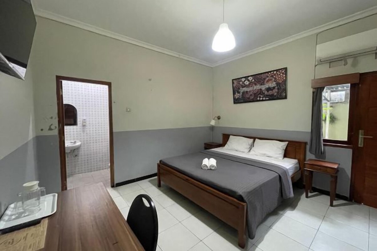 A bedroom with a bed, bedside table, and a small table with a chair. A bathroom is visible through an open door.