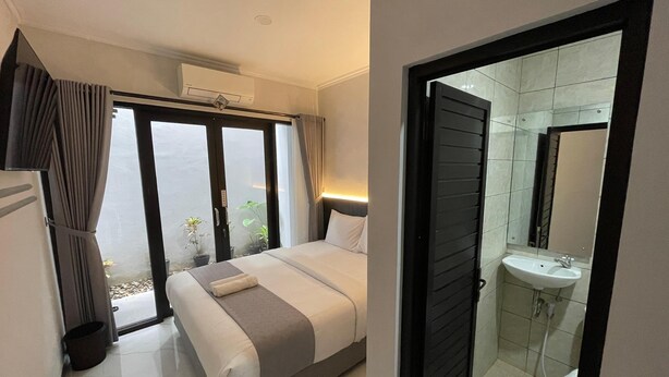 Una camera d'albergo con un letto, un bagno con lavandino e specchio, e una porta scorrevole in vetro che dà su un balcone con piante.