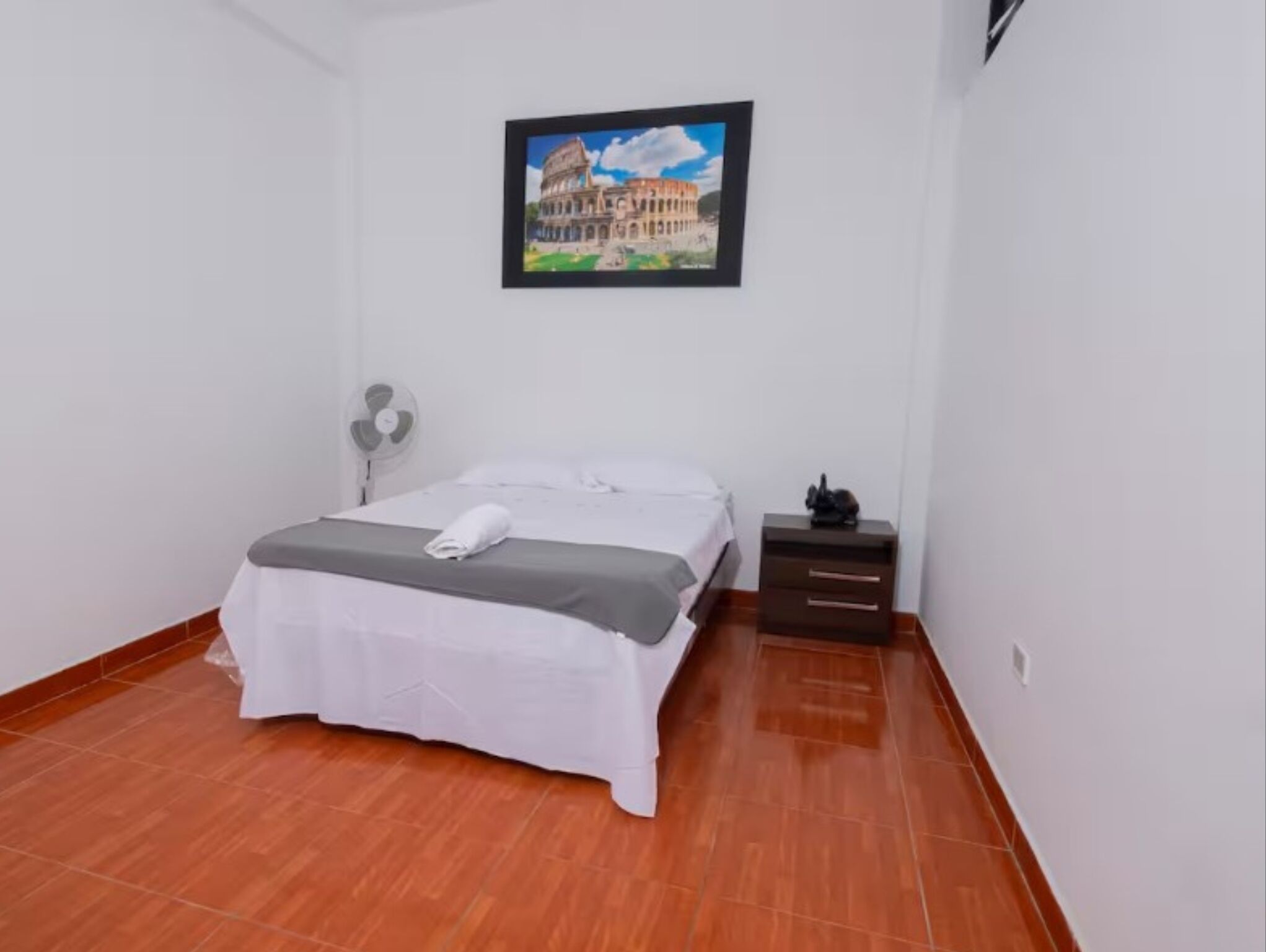A bedroom with a bed, a nightstand, a fan, and a framed picture of a building.