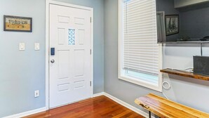 Una habitación con una puerta blanca, una ventana con persianas, un banco de madera y un estante con un altavoz.