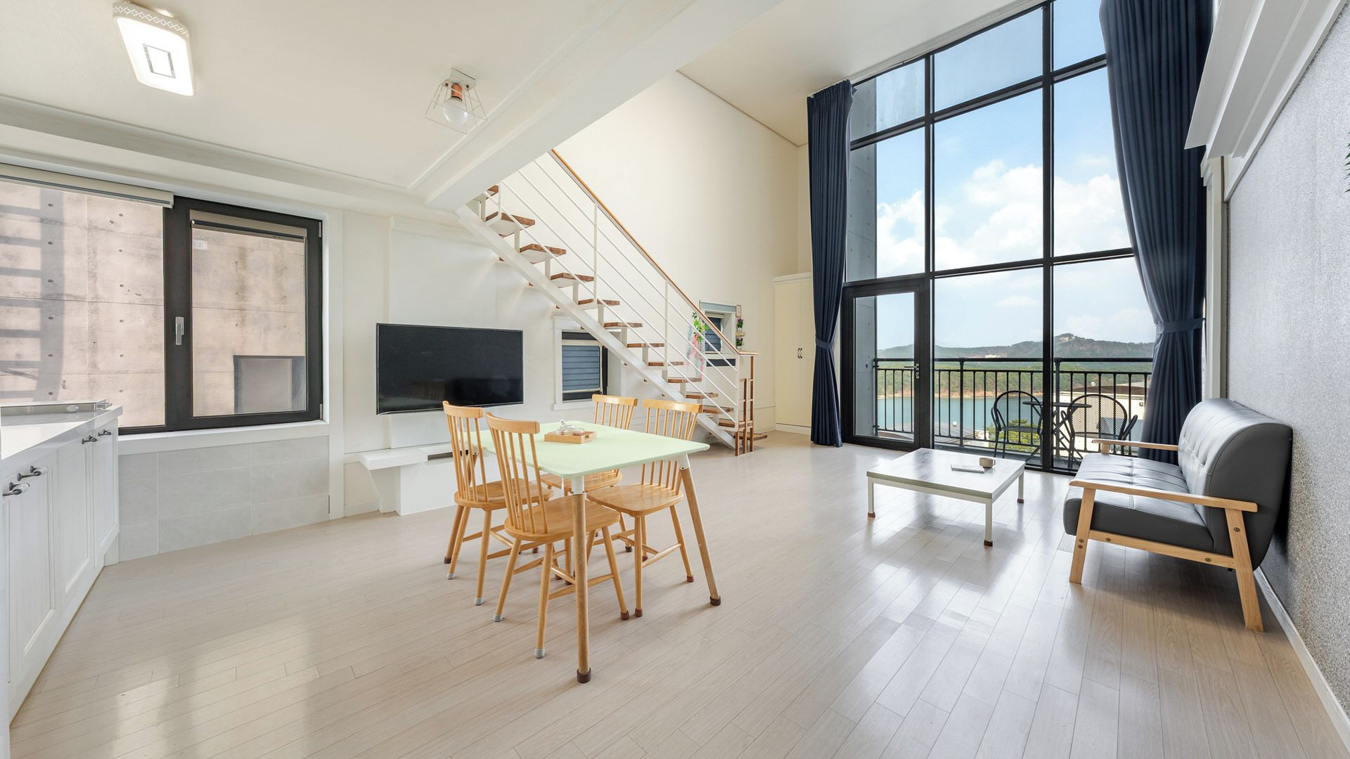 A spacious living area with a dining table, chairs, a sofa, a TV, and a staircase leading to a balcony with a view.