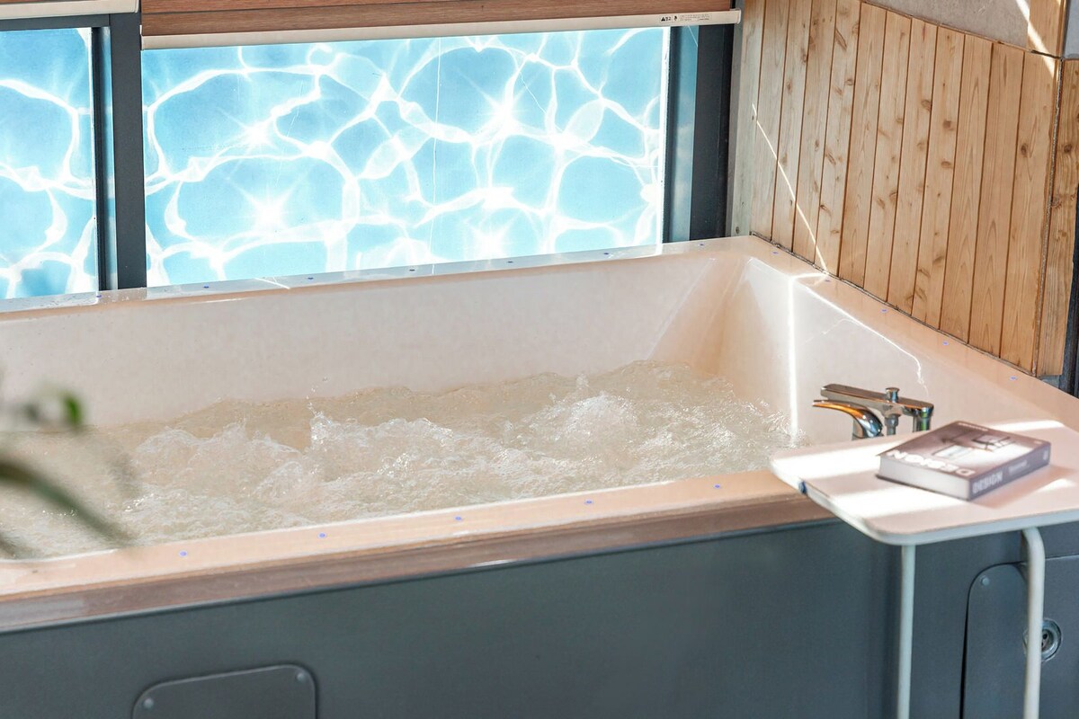 Eine Badewanne mit einer Wandpaneele aus Holz, ein Fenster mit blau schimmernder Aussicht und ein Buch am Rand.