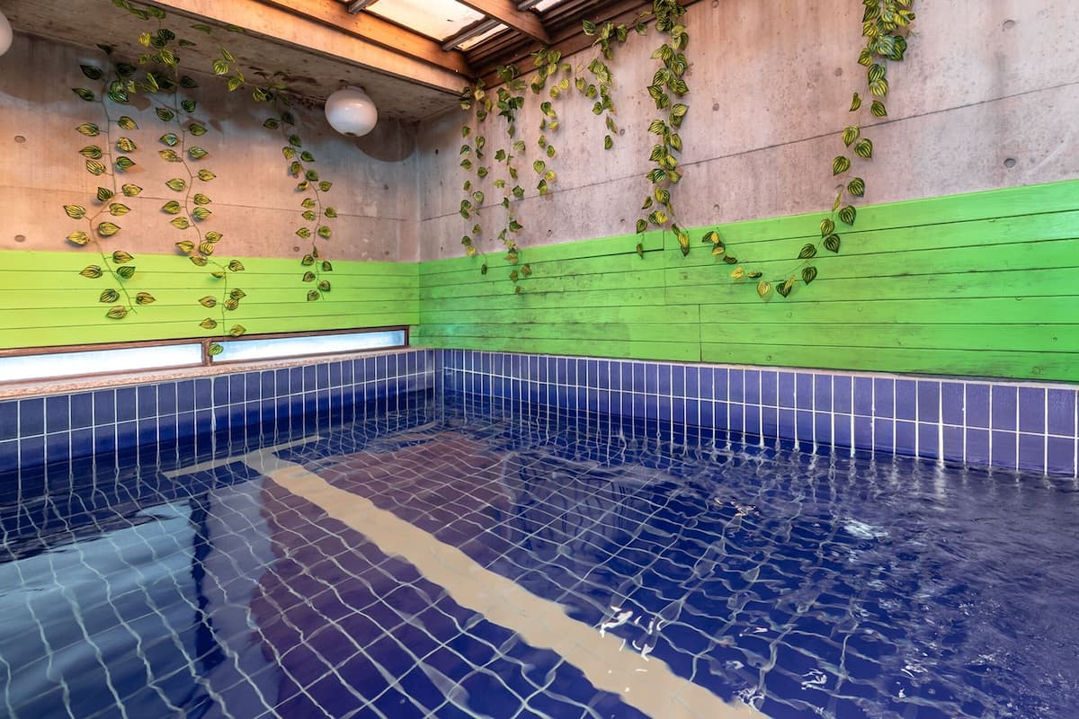 Una piscina con azulejos azules y paredes verdes, rodeada de suelo de baldosas.