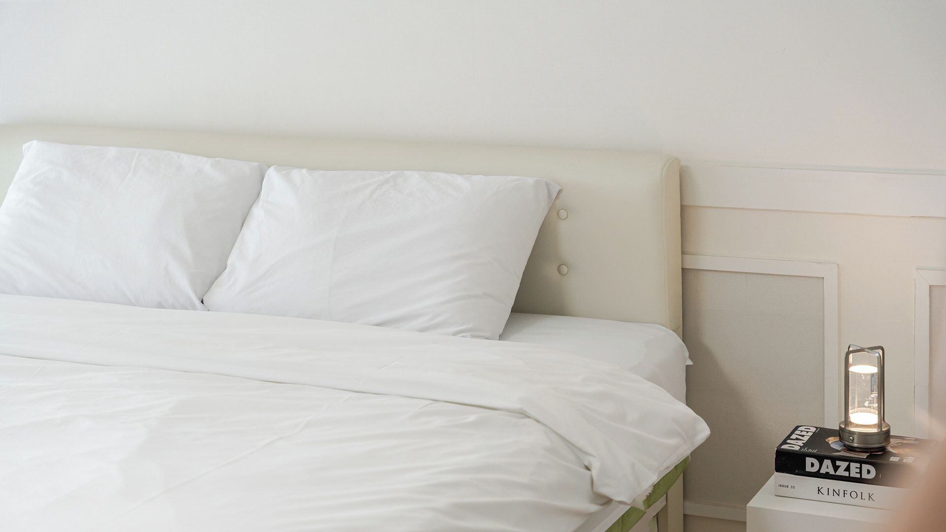 A neatly made bed with white bedding and pillows, a bedside table with a lamp, and a magazine titled 'DAZED' on the table.