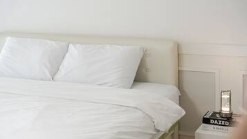 A neatly made bed with white bedding and pillows, a bedside table with a lamp, and a magazine titled 'DAZED' on the table.