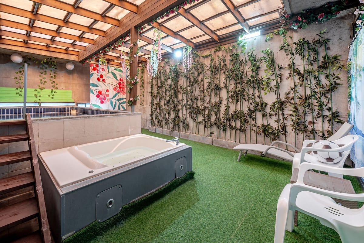 Un área de spa con una bañera grande, césped artificial verde, escaleras de madera y una pared decorada con plantas y flores colgantes.