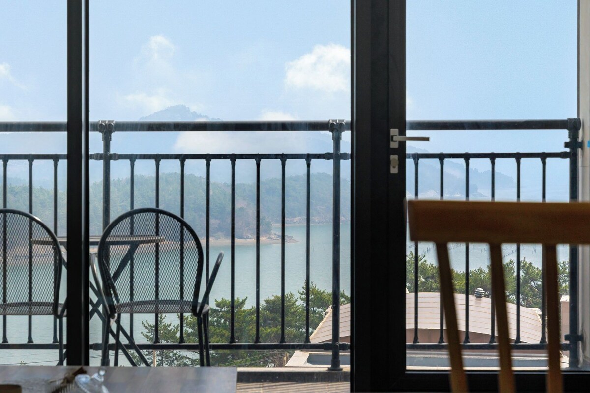Ein Balkon mit Blick auf ein Gewässer und einen fernen Berg. Zwei Stühle und ein Tisch befinden sich auf dem Balkon.