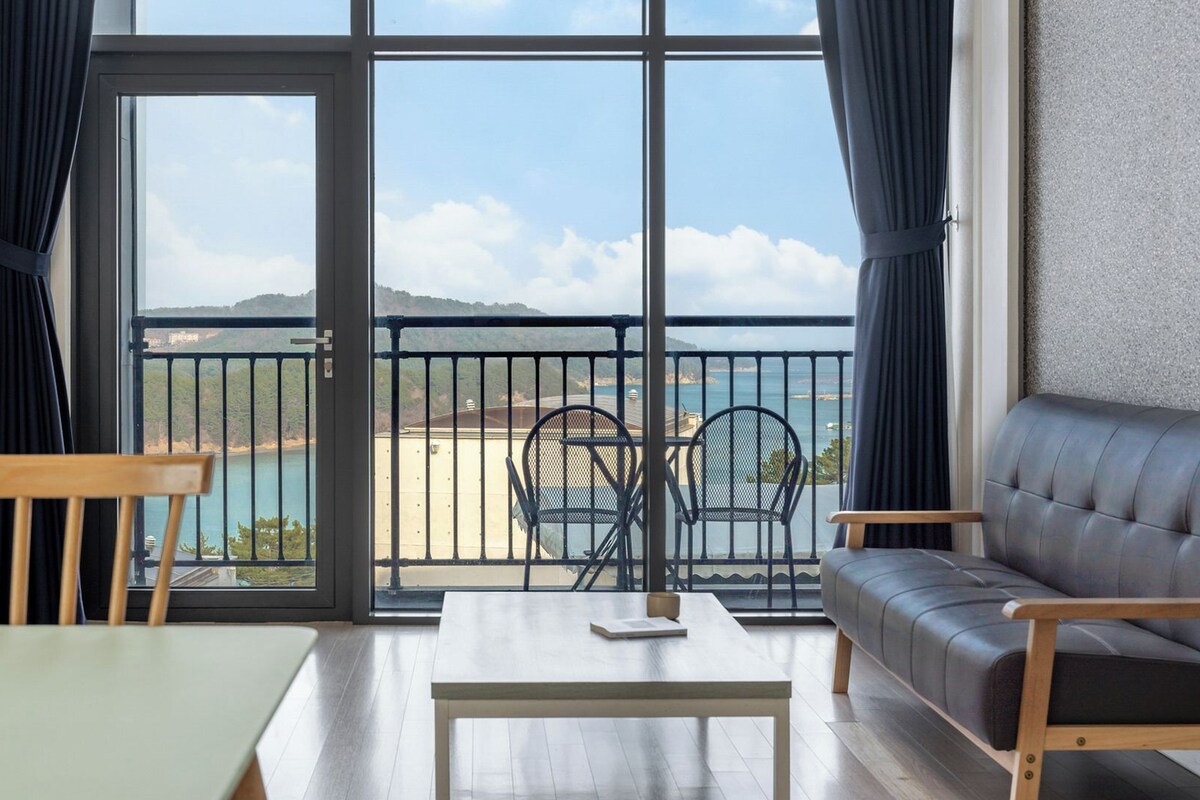 Ein Zimmer mit Balkon, einer Couch, einem Tisch und Stühlen mit Blick auf eine Küstenlandschaft.