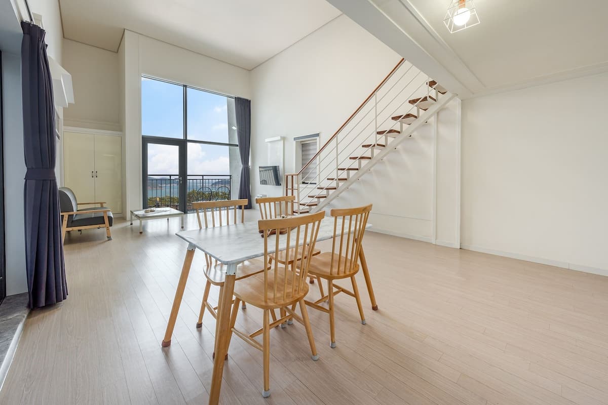 Una habitación amplia con suelo de madera, una mesa de comedor con sillas, una escalera y un gran ventanal con vistas al mar.