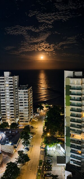 A nighttime view of a coastal urban area with high-rise buildings, a street with parked cars, and a visible moon.