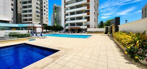 A swimming pool area with tiled flooring, lounge chairs, and a building with multiple balconies in the background.