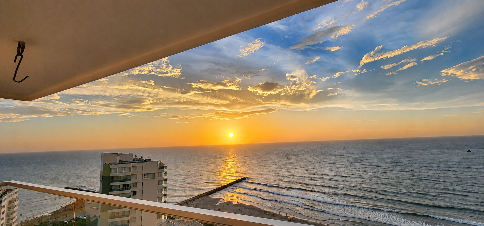 Une vue sur le coucher de soleil depuis un balcon donnant sur une plage, avec une jetée et des bâtiments au premier plan.